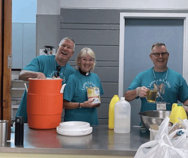Three people wearing blue "Loaves & Fishes" shirts prepare food and drinks in a kitchen, displaying camaraderie while participating in a community outreach program.