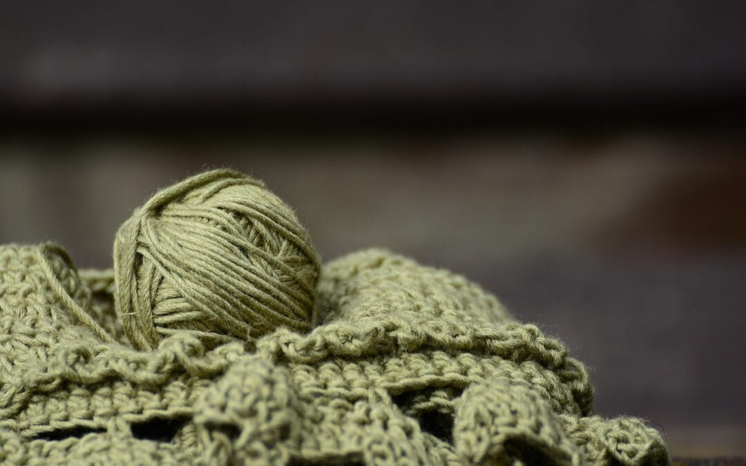 A ball of olive green yarn rests on a crocheted fabric piece, showcasing intricate patterns and soft textures.