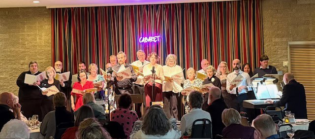 A group of people sings on stage with "CABARET" neon sign; audience seated at tables. Colorful curtain backdrop and piano visible.