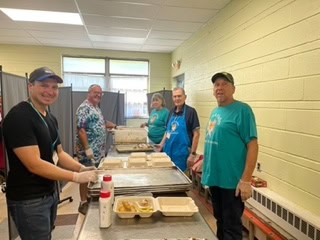 Five people working together in a kitchen setting preparing meals with trays and containers, wearing casual clothing and smiling at the camera.