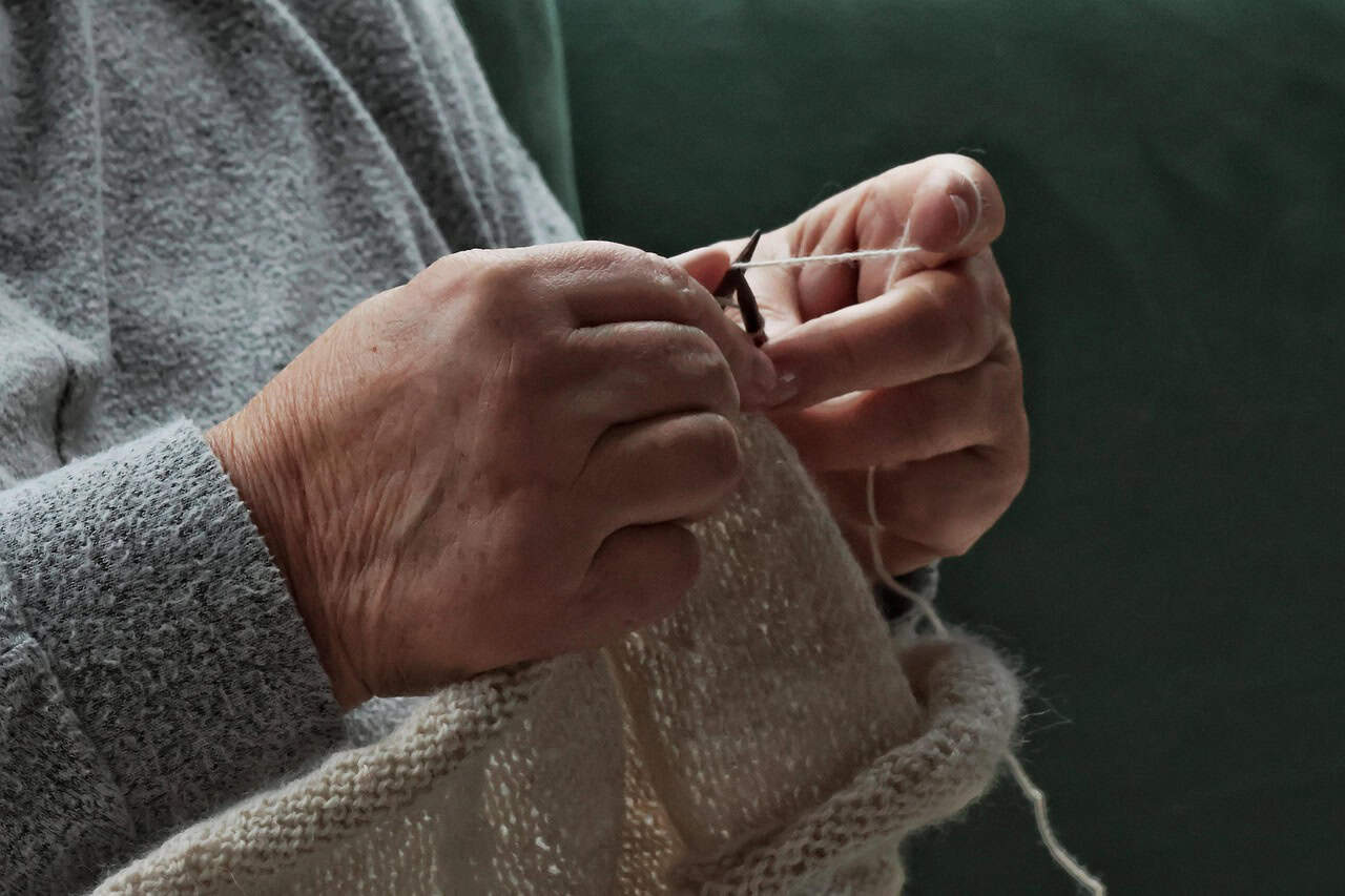 A person is knitting with white yarn, focusing on the intricate patterns. The background is a soft green, creating a calm atmosphere.