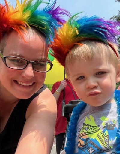 A person and child wear rainbow wigs, smiling outdoors. A vibrant atmosphere suggests a celebratory or festive event, with bright colors and a sunny day.