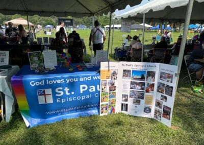 Outdoor event featuring St. Paul's Episcopal Church booth with informational display, colorful brochures, and people engaging under a tent in a grassy area.