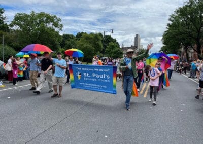 People march in a parade holding a "St. Paul's Episcopal Church" banner. Colorful umbrellas and a lively crowd fill the street.