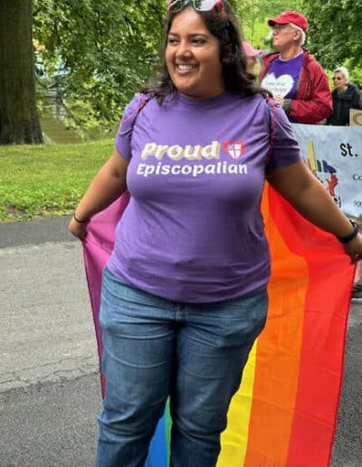 A person in a "Proud Episcopalian" shirt holds a rainbow flag at an outdoor event, surrounded by trees and a group of people.
