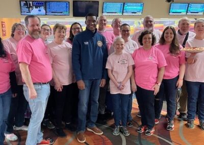 A group of people, mostly wearing pink shirts, stand together in a bowling alley with screens behind them, smiling for a group photo.